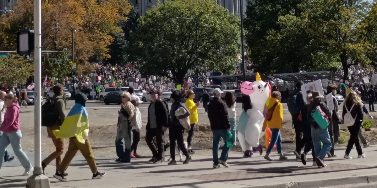 A group of protestors in inflatable costumes and holding protest signs cross the street into Civic Center Park minutes before the No Kings II protest in downtown Denver. 